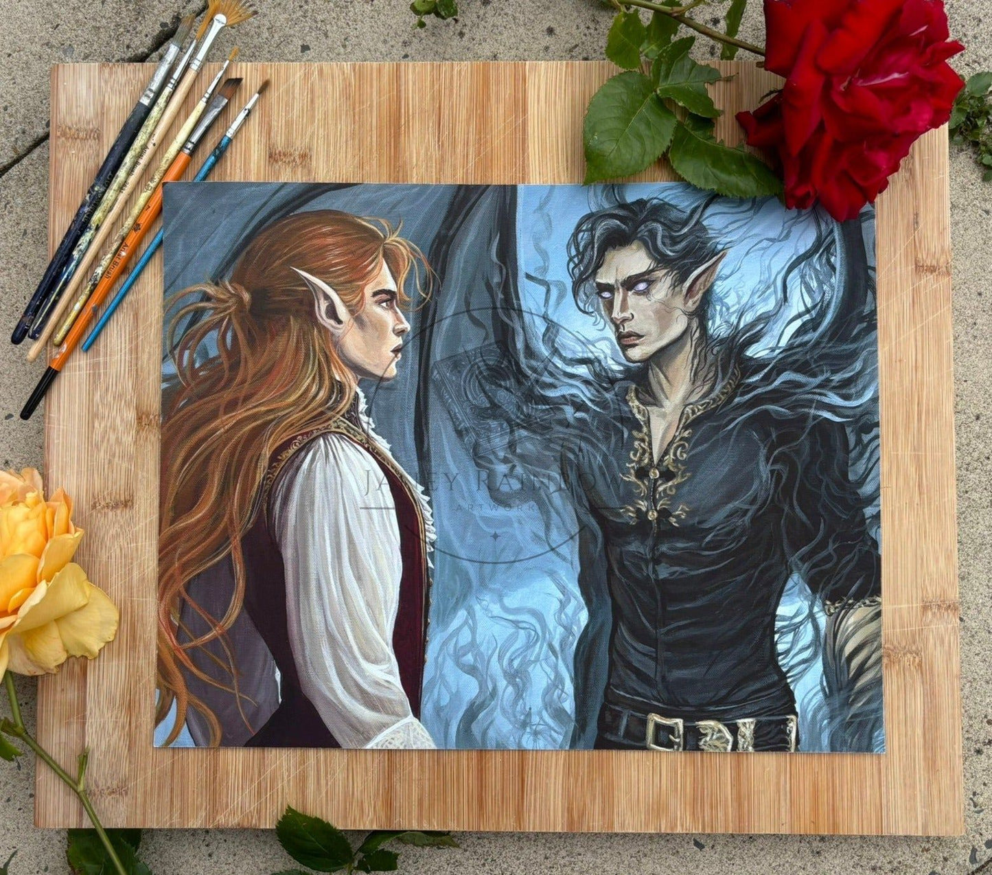 a print of a painting lies on a wooden board surrounded by roses and paintbrushes. The painting shows two faerie men staring at eachother intensely in a kind of show-down. The one on the left has long red hair and is wearing a crimson tunic with cream billowing sleeves. The man on the right is wearing dark colours with gold embroidery. He has large dark bat wings and violet eyes.