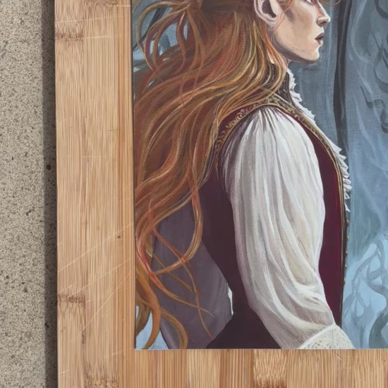 a print of a painting lies on a wooden board surrounded by roses and paintbrushes. The painting shows two faerie men staring at eachother intensely in a kind of show-down. The one on the left has long red hair and is wearing a crimson tunic with cream billowing sleeves. The man on the right is wearing dark colours with gold embroidery. He has large dark bat wings and violet eyes.