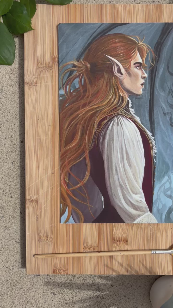 a print of a painting lies on a wooden board surrounded by roses and paintbrushes. The painting shows two faerie men staring at eachother intensely in a kind of show-down. The one on the left has long red hair and is wearing a crimson tunic with cream billowing sleeves. The man on the right is wearing dark colours with gold embroidery. He has large dark bat wings and violet eyes.